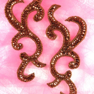 May include: Two gold-coloured, ornate, sequined embellishments in the shape of a stylised swirl on a pink background.
