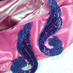 Könnte beinhalten: Zwei violette und blaue Perlenverzierungen in Form eines stilisierten Flügels. Die Verzierungen sind auf einen rosa Stoffhintergrund genäht.