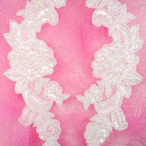 May include: Two white floral appliques with pearl-like embellishments on a pink background.