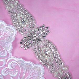Peut inclure: Une embellissement en perles argentées et blanches avec un motif floral. L'embellissement est sur un fond rose.
