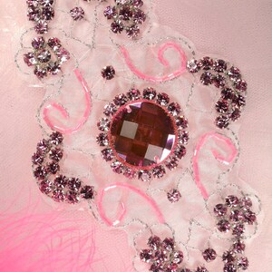 Może przedstawiać: Różowa i srebrna ozdoba z koralików z dużym różowym kryształem w środku. Ozdoba znajduje się na białym tle z różowym piórem w lewym dolnym rogu.