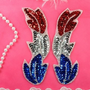 May include: Two sequined fabric patches in the shape of feathers. The feathers are red, white, and blue, resembling the colors of the American flag.