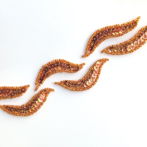 May include: Five orange sequined fabric appliques in the shape of a curved leaf. The appliques are arranged in a wave pattern.