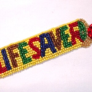 May include: A beaded bracelet with the word "LIFESAVER" spelled out in colorful beads. The bracelet is yellow with brown, blue, green, and red letters. A red, gem-like charm is attached to the end.