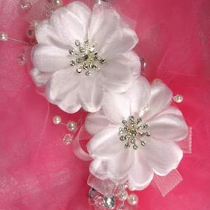 May include: Two white fabric flower hair accessories with rhinestone centers and pearl accents. The flowers are set against a pink background with sheer ribbons and additional embellishments. A delicate and elegant design.