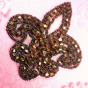 May include: A beaded floral applique in shades of bronze and gold, with a detailed, textured surface. The applique is set against a soft pink background, with a string of pearls in the lower right corner. The design is reminiscent of a stylized flower.