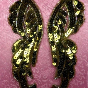 Puede incluir: Dos alas de lentejuelas doradas y negras con un diseño de plumas. Las alas están cosidas juntas en la parte superior y tienen aproximadamente 10 cm de altura.