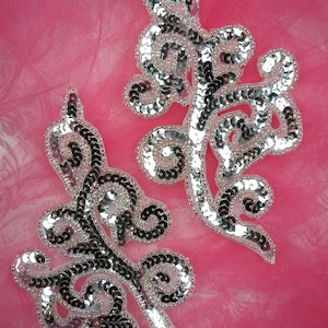 Puede incluir: Dos apliques de lentejuelas plateadas en forma de diseños florales estilizados sobre un fondo rosa.