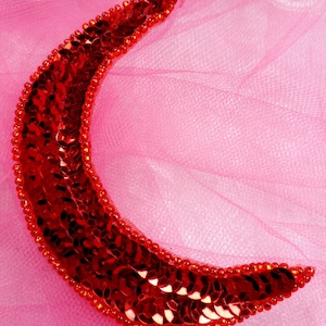 Op de afbeelding: Een rode halve maanvormige applicatie gemaakt van pailletten. De applicatie is op een roze tule achtergrond.