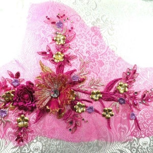 Peut inclure: Une applique en tissu rose avec des broderies florales dorées, roses et violettes et des paillettes. L'applique est sur un fond rose.