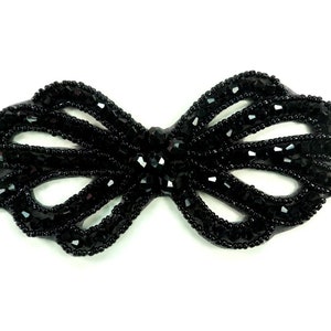 May include: A black bow-shaped applique crafted with faceted beads and small seed beads. The design features a central knot with two symmetrical loops on each side, creating an openwork pattern entirely covered in dark, reflective black beads.