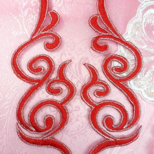 Puede incluir: Dos apliques de tela bordados en rojo y plata con un diseño de espiral. Los apliques están sobre un fondo de tela rosa.