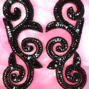 May include: Two black sequined embellishments in the shape of a stylized swirl design. The embellishments are on a pink fabric background.