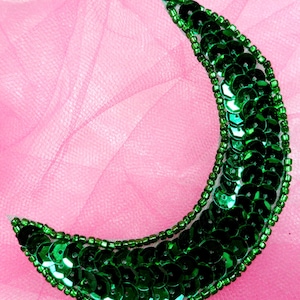 May include: A green crescent moon-shaped embellishment made of sequins and beads. The embellishment is on a pink background.