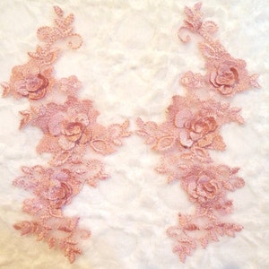 May include: Two pieces of delicate, rose-colored lace applique. Each applique features a floral design with detailed rose blossoms and leaves. The lace has a textured appearance, perfect for embellishing garments or crafts.