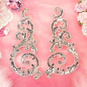 Könnte beinhalten: Zwei silberne Pailletten-Stoffapplikationen in einem zarten Blumenmuster. Die Applikationen befinden sich auf einem rosa Hintergrund.