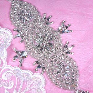 Peut inclure: Applique en strass argenté avec un motif floral. L'applique est allongée avec une forme ovale centrale et ornée de nombreux strass transparents. Le fond est un tissu rose clair, et une bordure en dentelle blanche est visible à gauche.