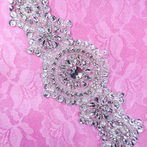 Peut inclure: Une embellissement en perles argentées et blanches avec un grand motif circulaire en strass au centre. L'embellissement est sur un fond de tissu rose.