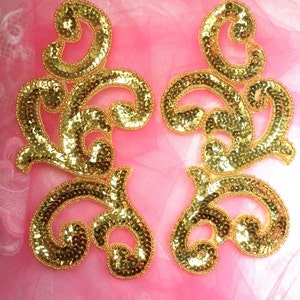 May include: Four gold sequined appliques in a swirling, ornate design. Each applique is made of gold sequins and outlined with gold thread. The appliques are arranged on a pink, sheer fabric background.