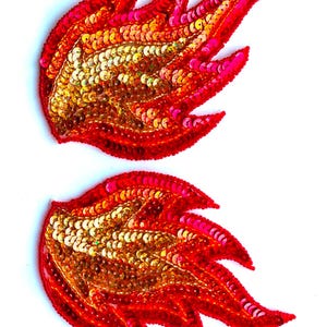 Könnte beinhalten: Zwei dekorative Flügel-Applikationen mit Pailletten. Die Flügel sind hauptsächlich rot mit goldenen und rosa Akzenten. Die Pailletten sind dicht gepackt und erzeugen einen schimmernden Effekt. Ideal zum Verzieren von Kleidung oder Accessoires.