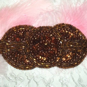 May include: A brown beaded embellishment with a circular design. The embellishment is made of small, round beads and has a three-dimensional shape.