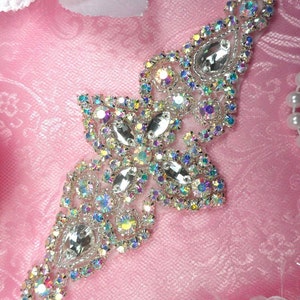 May include: A sparkling, ornate rhinestone applique in a cross-like design. The applique features large, clear teardrop-shaped rhinestones and smaller, iridescent, colorful rhinestones. The piece is set on a pink fabric background.