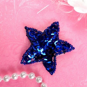 May include: A blue star-shaped embellishment made of sequins on a pink background. A string of white pearls is in the lower left corner of the image.