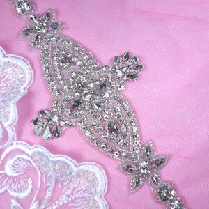 Peut inclure: Une embellissement en strass argenté avec un motif floral. L'embellissement est sur un fond rose.