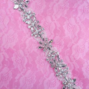 Peut inclure: Une garniture de perles argentées avec des strass clairs disposés en motif floral. La garniture est sur un fond de tissu rose.