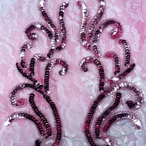Puede incluir: Dos apliques simétricos con un diseño floral en espiral. Los apliques están hechos de lentejuelas rosas brillantes y están delineados con cuentas plateadas. Se colocan sobre un fondo de encaje rosa.