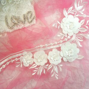 Peut inclure: Garniture en dentelle blanche avec un motif floral et des accents de perles. La garniture présente des fleurs 3D en relief et des feuilles délicates. La garniture est sur un fond rose.
