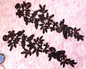 May include: Two black lace appliques with floral and leaf designs. The appliques are symmetrical, with a delicate, openwork pattern. The lace is detailed with small flowers and leaves, creating an elegant and intricate design. The appliques are set against a pink lace background.