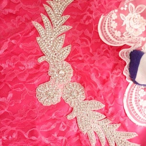 Peut inclure: Une applique en strass argentée avec un motif floral. L'applique est sur un tissu en dentelle rose.