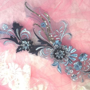 Peut inclure: Une applique florale bleue et noire avec des paillettes. L'applique est sur un fond de tulle rose.