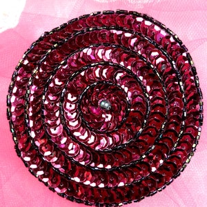 May include: A close-up of a circular, burgundy-colored embellishment made of sequins. The sequins are arranged in a spiral pattern, creating a swirling effect.