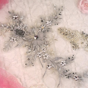 Peut inclure: Deux appliques florales argentées avec des perles et des strass. Les appliques sont sur un fond blanc.