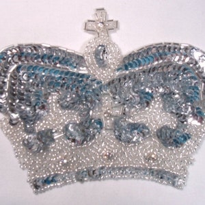 May include: A silver and blue sequined crown with a cross on top. The crown is made of small, round sequins and beads.