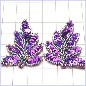 May include: Two purple sequined leaf-shaped embellishments. The sequins are iridescent and have a slightly raised texture. The embellishments are perfect for adding a touch of sparkle to any craft project.