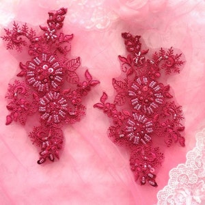 May include: Two burgundy floral lace appliques with beaded embellishments. The appliques are on a pink background.