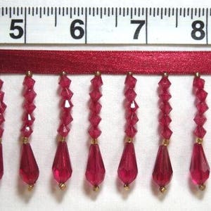 May include: Close-up of a decorative trim featuring a burgundy ribbon with hanging ruby red faceted beads. The beads are teardrop-shaped and connected with gold-colored accents. A ruler in inches is visible above the trim.