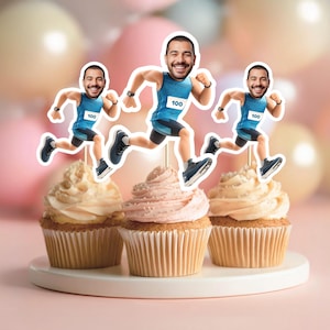 Puede incluir: Adornos para cupcakes con un corredor sonriente, con una camiseta sin mangas azul con el número 100, pantalones cortos negros y zapatillas para correr. Los adornos están colocados sobre cupcakes con glaseado blanco y rosa. El fondo tiene globos rosas y beige.