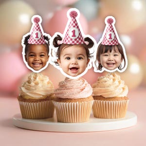 May include: Three cupcakes with smiling baby faces wearing party hats with the number 1. The cupcakes have vanilla and pink frosting, and are on a white plate. Balloons in the background.