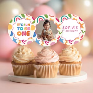 May include: Three cupcakes with decorative toppers. One topper says "It's Fun To Be One", another shows a baby with a teddy bear, and the third says "Sofia's 1st Birthday". The cupcakes have vanilla and pink icing.