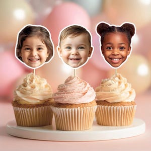 May include: Three cupcakes topped with smiling children's faces. The cupcakes have light-coloured frosting and are on a white plate. Pink and beige balloons are in the background.