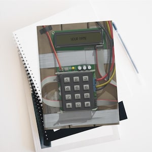 Pode incluir: Um caderno com um design impresso que se assemelha a uma bomba. O design inclui um teclado, fios e um visor digital que diz "YOUR NAME". O caderno está em cima de outros cadernos e uma caneta.