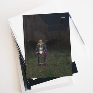 Pode incluir: Um caderno de espiral com uma ilustração na capa de um personagem fantástico de pele amarela, vestindo uma túnica branca e verde com luvas e botas roxas, em frente a uma estrutura de pedra escura com duas grandes figuras sombrias.