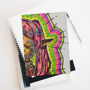 May include: A spiral-bound notebook featuring a colorful illustration of a person with pink hair and a rainbow-colored halo. The artwork has a unique, hand-drawn style with bold lines and vibrant colors. The notebook is resting on top of other notebooks.