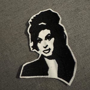 May include: A black and white embroidered patch depicting a woman with dark hair. The patch has a white border and is likely an iron-on or sew-on patch for clothing or accessories.