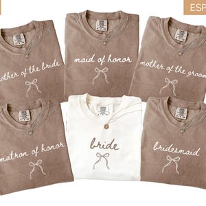 May include: An assortment of t-shirts in ivory and espresso shades, featuring white script text: "mother of the bride", "maid of honor", "mother of the groom", "matron of honor", "bride", and "bridesmaid". Each shirt includes a bow graphic.