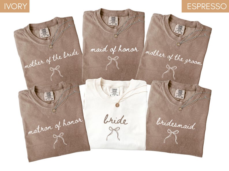 May include: An assortment of t-shirts in ivory and espresso shades, featuring white script text: "mother of the bride", "maid of honor", "mother of the groom", "matron of honor", "bride", and "bridesmaid". Each shirt includes a bow graphic.